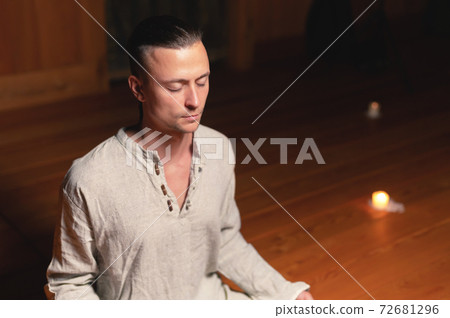 A man in clothes for practice and meditation sits in a lotus pose and holds red rosary to concentrate attention in a wooden room with dim light A man in clothes for practice and meditation sits in a lotus pose and holds red rosary to concentrate attention in a wooden room with dim light 72681296