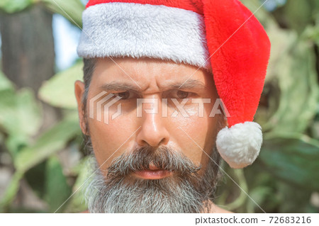 Close-up portrait of a tanned, bearded caucasian man in a Santa Claus hat standing in front of cacti on the beach.  72683216