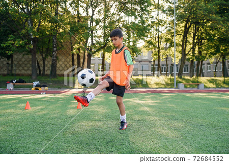 Motivated teen football player stuffs soccer ball on leg. Practicing sport exercises at artificial stadium. Motivated teen football player stuffs soccer ball on leg. Practicing sport exercises at artificial stadium. 72684552