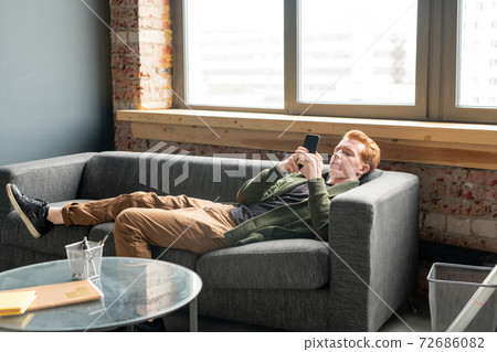 Young restful male specialist using smartphone while relaxing on soft couch 72686082