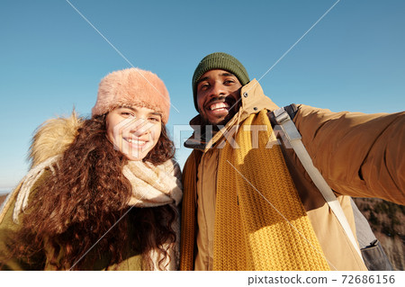 Happy young intercultural dates in winterwear making selfie against blue sky 72686156