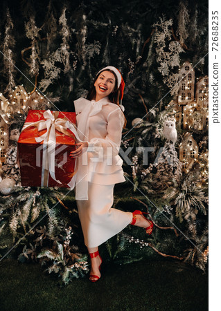 a girl in a Santa hat with a big Christmas gift in her hands on a fairy-tale island.Smiling woman in white clothes on the background of Christmas trees and small houses a girl in a Santa hat with a big Christmas gift in her hands on a fairy-tale island.Smiling woman in white clothes on the background of Christmas trees and small houses 72688235