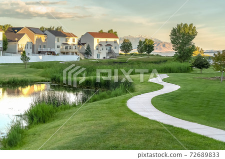 Pond and pathway on a scenic park in front of sunlit houses under cloudy sky 72689833