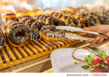 Woman using tongs picking tasty frosted chocolate donut at bakery Woman using tongs picking tasty frosted chocolate donut at bakery 72692399
