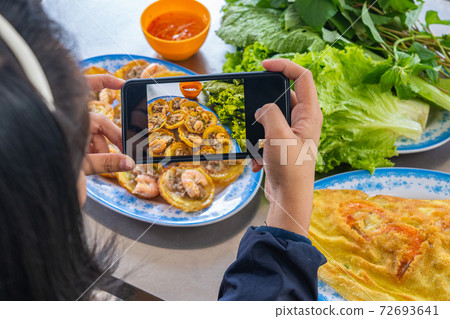 Young woman taking photo by smartphone while eating Banh Khot 72693641