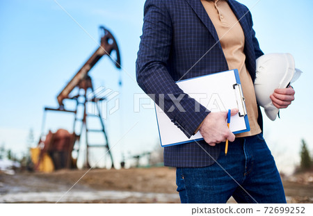 Oil worker holding white safety helmet and clipboard. 72699252