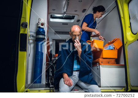 Injured man in a blanket breathing through an oxygen mask in an ambulance car, nurse packing medical equipment in the background 72700252