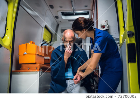 Man sitting with an oxygen mask in an ambulance, woman in a medical uniform is checking his pulse Man sitting with an oxygen mask in an ambulance, woman in a medical uniform is checking his pulse 72700302