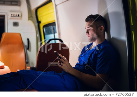 Young paramedic sitting with his smartphone in the ambulance car Young paramedic sitting with his smartphone in the ambulance car 72700527