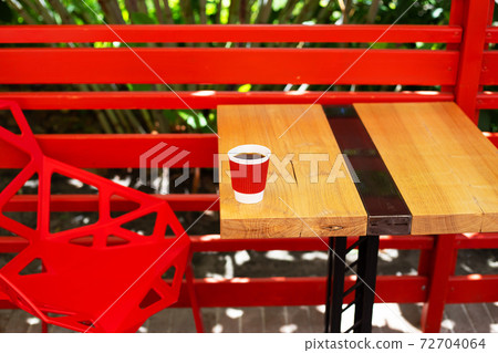 A wooden table on which there is a red plastic cup with coffee. Summer beautiful terrace in greenery 72704064