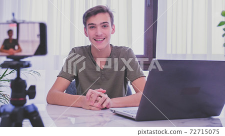 Man blogger videotapes his vlog at home. Teenage boy recording his video blog. Young man speaks in front of a video camera for his blog channel. Vlogger makes online streaming using smartphone 72715075