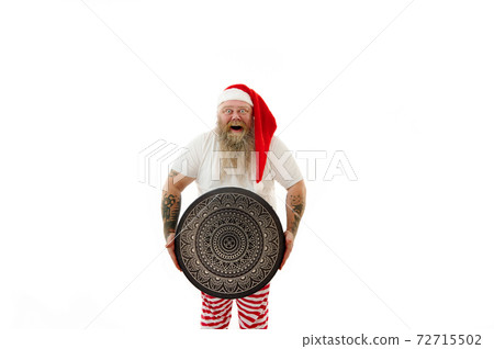 An overweigh happy and joyful man shaman with tattooed arms in Santa Claus hat holding a shaman tambourine with beautiful luxury mandala. Isolated portrait on a white background An overweigh happy and joyful man shaman with tattooed arms in Santa Claus hat holding a shaman tambourine with beautiful luxury mandala. Isolated portrait on a white background 72715502