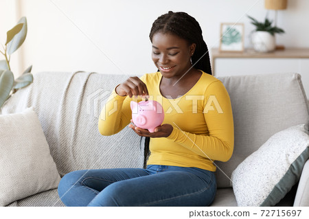Black woman putting coin in piggy bank, sitting on couch at home Black woman putting coin in piggy bank, sitting on couch at home 72715697