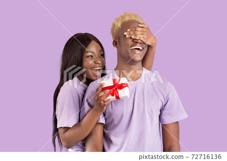 Pretty black woman covering her boyfriend's eyes, surprising him with Valentine's gift on lilac studio background 72716136