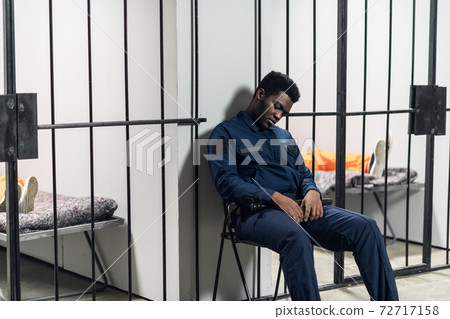 A strong black prison guard guards the cells with prisoners in the prison corridor A strong black prison guard guards the cells with prisoners in the prison corridor 72717158