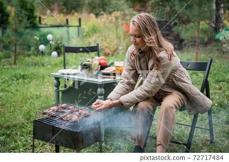 An optimistic young girl is preparing a barbecue for the whole family in the courtyard of the family mansion. Autumn holidays in self-isolation. An optimistic young girl is preparing a barbecue for the whole family in the courtyard of the family mansion. Autumn holidays in self-isolation. 72717484