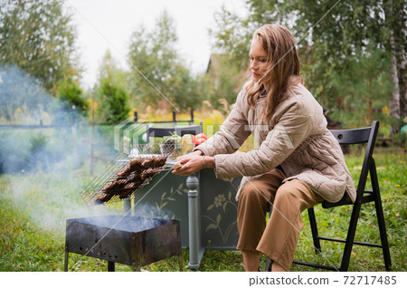 Bored in quarantine, a young woman roasts a barbecue on the grill. A table with vegetables, snacks and beer is set nearby. 72717485