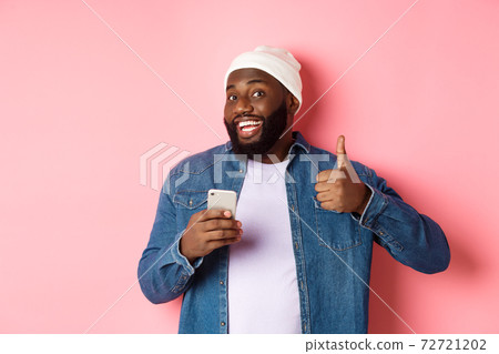 Technology and online shopping concept. Satisfied Black man smiling, using mobile phone and showing thumb-up in approval, making order, standing over pink background Technology and online shopping concept. Satisfied Black man smiling, using mobile phone and showing thumb-up in approval, making order, standing over pink background 72721202