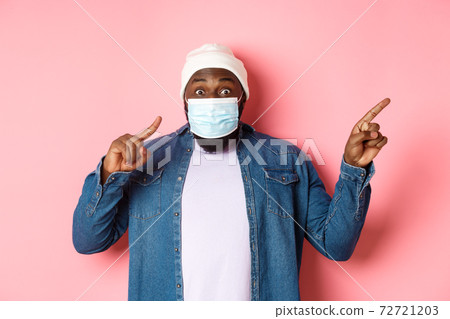 Covid-19, lifestyle and lockdown concept. Impressed Black man in face mask showing announcement, pointing right at promo and staring at camera amazed, pink background Covid-19, lifestyle and lockdown concept. Impressed Black man in face mask showing announcement, pointing right at promo and staring at camera amazed, pink background 72721203