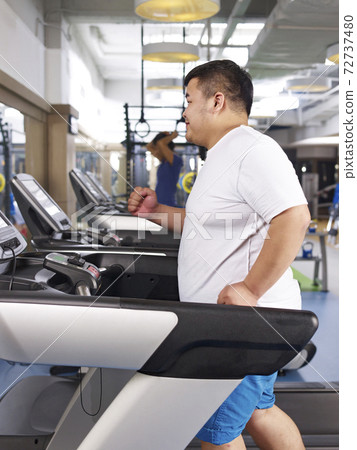 an overweight young man running on treadmill in fitness center 72737480