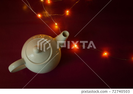 White ceramic kettle and Christmas lights on burgundy background White ceramic kettle and Christmas lights on burgundy background 72740503