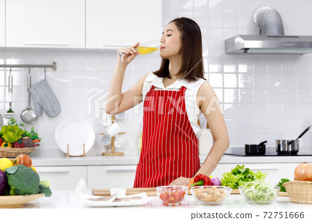 Asian woman in red apron standing in the kitchen and drinking glass of champagne. Concept woman cooking at home. Asian woman in red apron standing in the kitchen and drinking glass of champagne. Concept woman cooking at home. 72751666