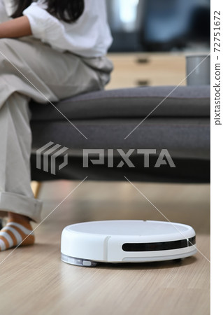 Robotic vacuum cleaner cleaning the living room and woman sitting on sofa at home. Robotic vacuum cleaner cleaning the living room and woman sitting on sofa at home. 72751672