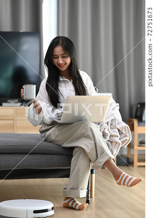 Happy woman sitting on sofa working with laptop and drinking coffee while robot vacuum cleaning floor at home. Happy woman sitting on sofa working with laptop and drinking coffee while robot vacuum cleaning floor at home. 72751673