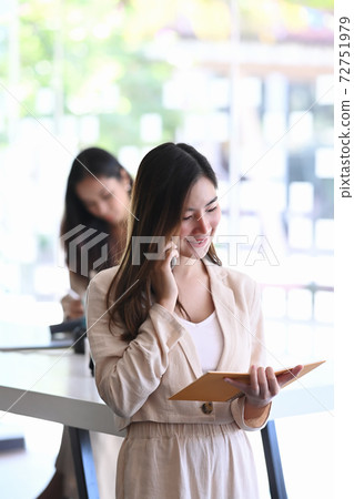 Portrait of young businesswoman talking on mobile phone and looking information in notebook while standing in meeting room. 72751979