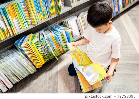 Asian little child sit on the floor,reading a cartoon from a fable book,happy kid boy enjoy in school library,excited kindergarten student looking at picture and color from fable book in a bookstore 72758393