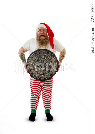 Isolated full-length portrait on a white background of an overweigh funny shaman with tattooed arms in Santa Claus hat holding a shaman tambourine with beautiful luxury mandala ornament. Isolated full-length portrait on a white background of an overweigh funny shaman with tattooed arms in Santa Claus hat holding a shaman tambourine with beautiful luxury mandala ornament. 72766406