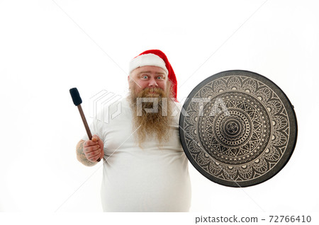 Isolated portrait on a white background of an overweigh smiling man shaman with tattooed arms in Santa Claus hat holding a shaman tambourine expressing emotion. Isolated portrait on a white background of an overweigh smiling man shaman with tattooed arms in Santa Claus hat holding a shaman tambourine expressing emotion. 72766410
