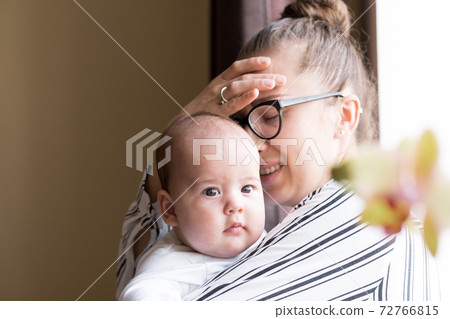 Childhood, motherhood, illness family concepts - upset, tired nervous business woman Lady mother with headache holding infant child newborn baby in her hands stands near window at home indoors 72766815
