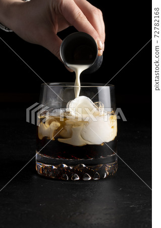 A girl bartender prepares a White Russian cocktail and pours cream into it. Cold alcoholic cocktail of cream, coffee liqueur, vodka and ice in a transparent glass on a black background. 72782168