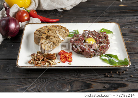 Beef tartare. Cold appetizer from raw marinated meat with quail egg, bread, chilli and pecan in a ceramic plate on a wooden kitchen table. 72782214