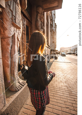 A girl with blond hair in a checkered dress and a black leather jacket is standing backwards and looking forward into the street in the old town. Backlight. Egyptian House in St. Petersburg, Russia A girl with blond hair in a checkered dress and a black leather jacket is standing backwards and looking forward into the street in the old town. Backlight. Egyptian House in St. Petersburg, Russia 72786116
