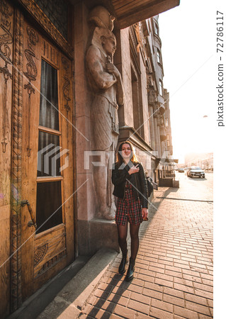 Attractive girl with blond hair in a dress in a cage and a black leather jacket in the sunlight goes forward to the camera. behind the Egyptian house with the statues of the pharaohs at the entrance Attractive girl with blond hair in a dress in a cage and a black leather jacket in the sunlight goes forward to the camera. behind the Egyptian house with the statues of the pharaohs at the entrance 72786117