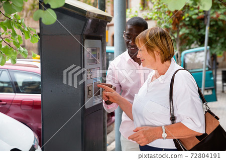 Polite intelligent African man helping middle aged woman to buy ticket in parking meter on summer city street Polite intelligent African man helping middle aged woman to buy ticket in parking meter on summer city street 72804191