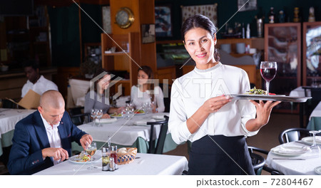 Hospitable waitress with serving tray meeting guests in restaurant Hospitable waitress with serving tray meeting guests in restaurant 72804467