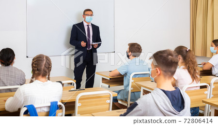 Teacher in protective mask with notebook is giving interesting lecture for students in classroom 72806414