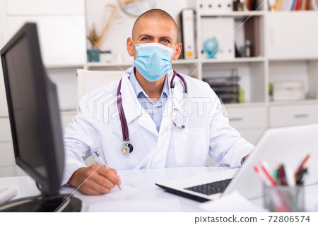 Man doctor in medical mask is working at laptop in clinic Man doctor in medical mask is working at laptop in clinic 72806574