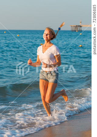 Smiling adult woman in white T-shirt is jogging on the beach Smiling adult woman in white T-shirt is jogging on the beach 72806885
