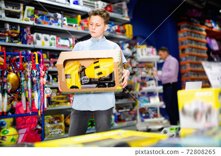Serious teen boy looking narrowly at yellow plastic car in children toy store 72808265