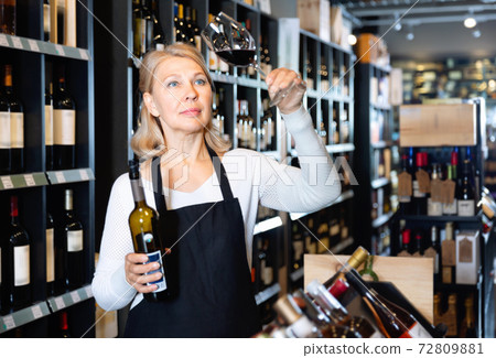 Focused mature woman wine producer inspecting quality of wine in wineshop on background with shelves of wine bottles Focused mature woman wine producer inspecting quality of wine in wineshop on background with shelves of wine bottles 72809881