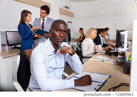 Puzzled african businessman sitting at desk in open plan office, thinking about business strategy Puzzled african businessman sitting at desk in open plan office, thinking about business strategy 72809901