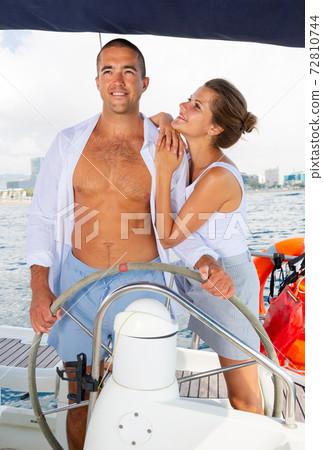 Couple standing at yacht wheel along coast of Barcelona Couple standing at yacht wheel along coast of Barcelona 72810744