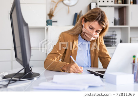 Business woman in protective medical mask is working with project behind laptop Business woman in protective medical mask is working with project behind laptop 72811369