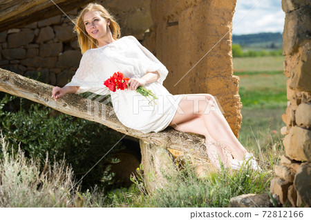 Young female in white dress posing on tree trunk with poppy flowers in hands Young female in white dress posing on tree trunk with poppy flowers in hands 72812166