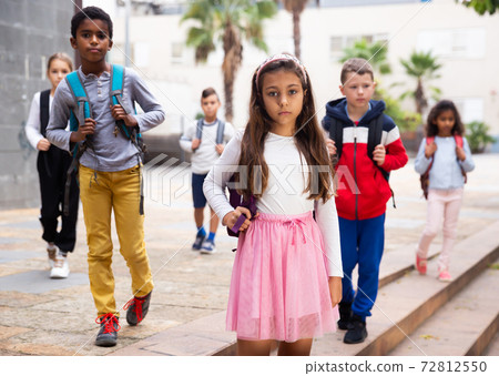 Positive schoolgirl standing near school, children on background Positive schoolgirl standing near school, children on background 72812550