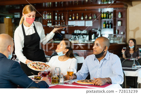 Waitress in protective mask serving delicious pizza to friends in restaurant Waitress in protective mask serving delicious pizza to friends in restaurant 72813239